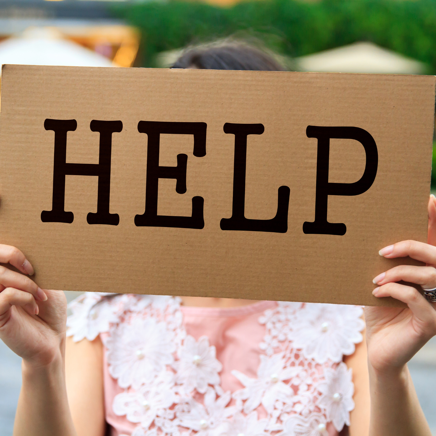 A woman is holding up a "HELP" sign on cardboard. the background is blurred. her nails are well manicured and she's wearing a soft lacy blouse.
