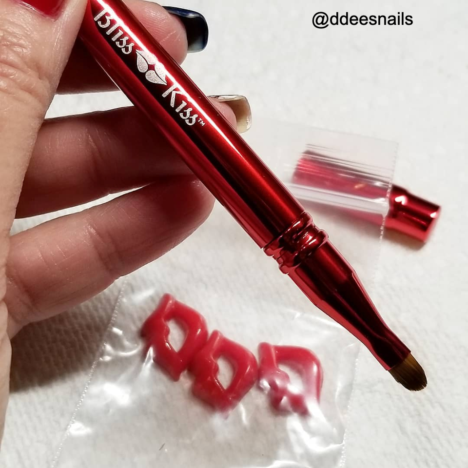 @ ddeesnails
Image shows a hand holding a red Bliss Kiss nail brush. the nail brush is elegant and metallic.
in the background their is cute lip shaped designs that could be placed on a nail.