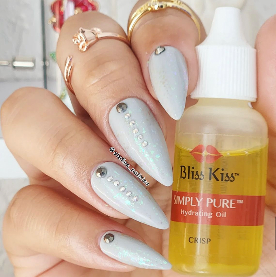 @ stefies_nailzies
Image shows a hand with well polished stiletto nails. the nail polish is white with rhinestones going down the center. The hand is softly holding a bottle of Bliss Kiss simply pure hydrating oil in the scent Crisp. 